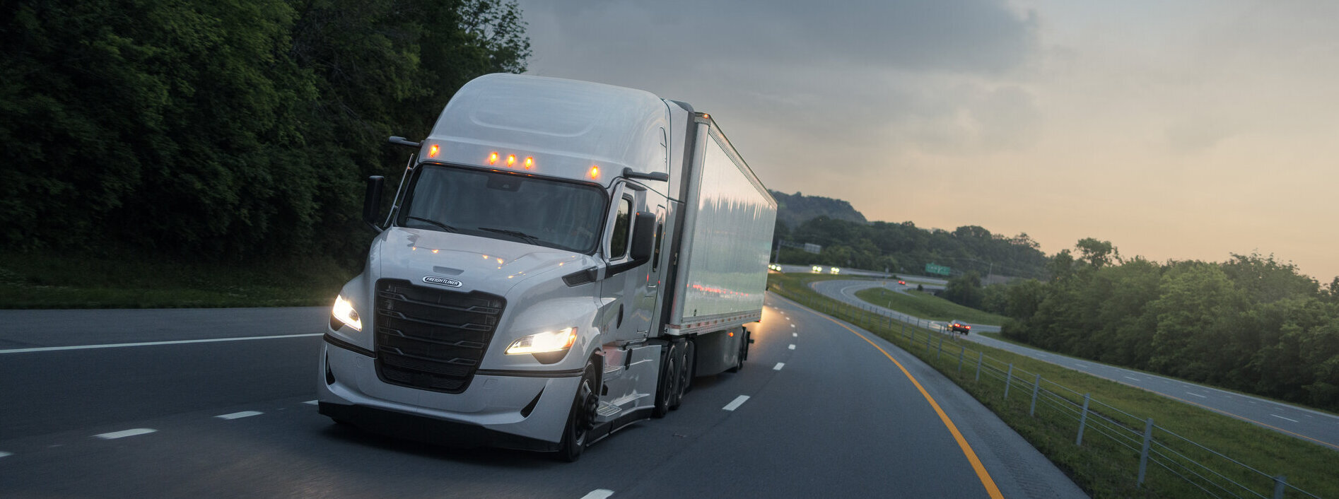 A white semi truck with a plain, unbranded trailer running the highway at dusk
